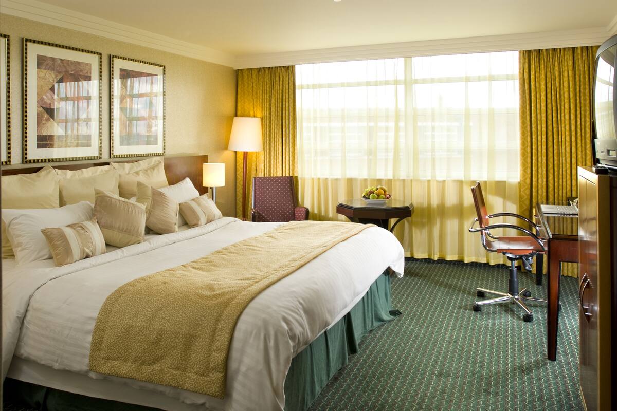 A hotel room with a large bed, a desk with a chair, a small table with a fruit bowl, and a television.