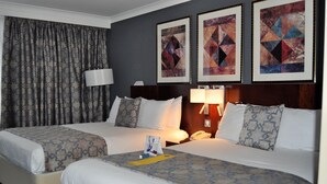 A hotel room with two beds, a large window with curtains, a desk with a lamp, and artwork on the wall.