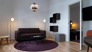 Ein modernes Wohnzimmer mit einem dunklen Sofa, einem runden Couchtisch und einem violetten Teppich. Zwei Stehlampen, ein an der Wand montierter Fernseher und ein kleiner Beistelltisch sind ebenfalls vorhanden.