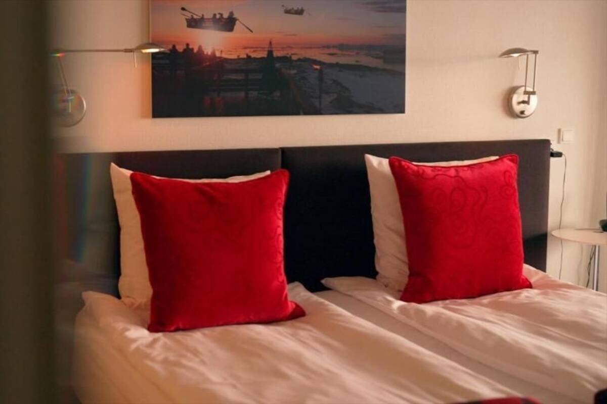 Una cama con almohadas blancas y rojas, un cabecero oscuro y un cuadro con una escena de atardecer en la pared.
