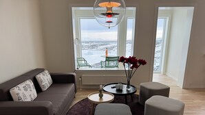Ein modernes Wohnzimmer mit einer Couch, Hocker, einem Couchtisch und Blick auf eine verschneite Landschaft durch große Fenster.