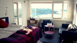 A hotel room with a bed, a desk, a chair, and a sofa. There is a view of the beach through the window. - Hotel Arctic (Ilulissat)