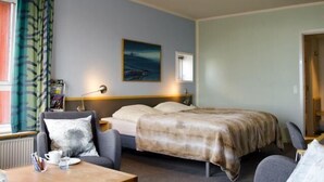 A hotel room with a bed, a desk with a chair, a window with curtains, a painting on the wall, and a radiator. - Hotel Arctic (Ilulissat)