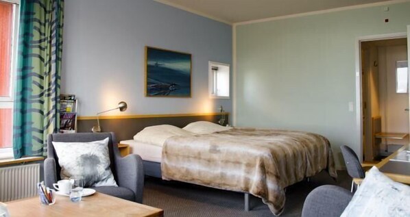A hotel room with a bed, a desk with a chair, a window with curtains, a painting on the wall, and a radiator. - Hotel Arctic (Ilulissat)