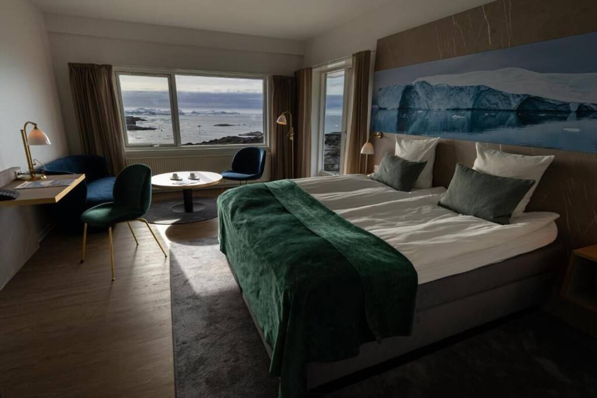 Habitación de hotel con una cama grande, un escritorio con silla, una mesita y vistas al océano a través de amplias ventanas.