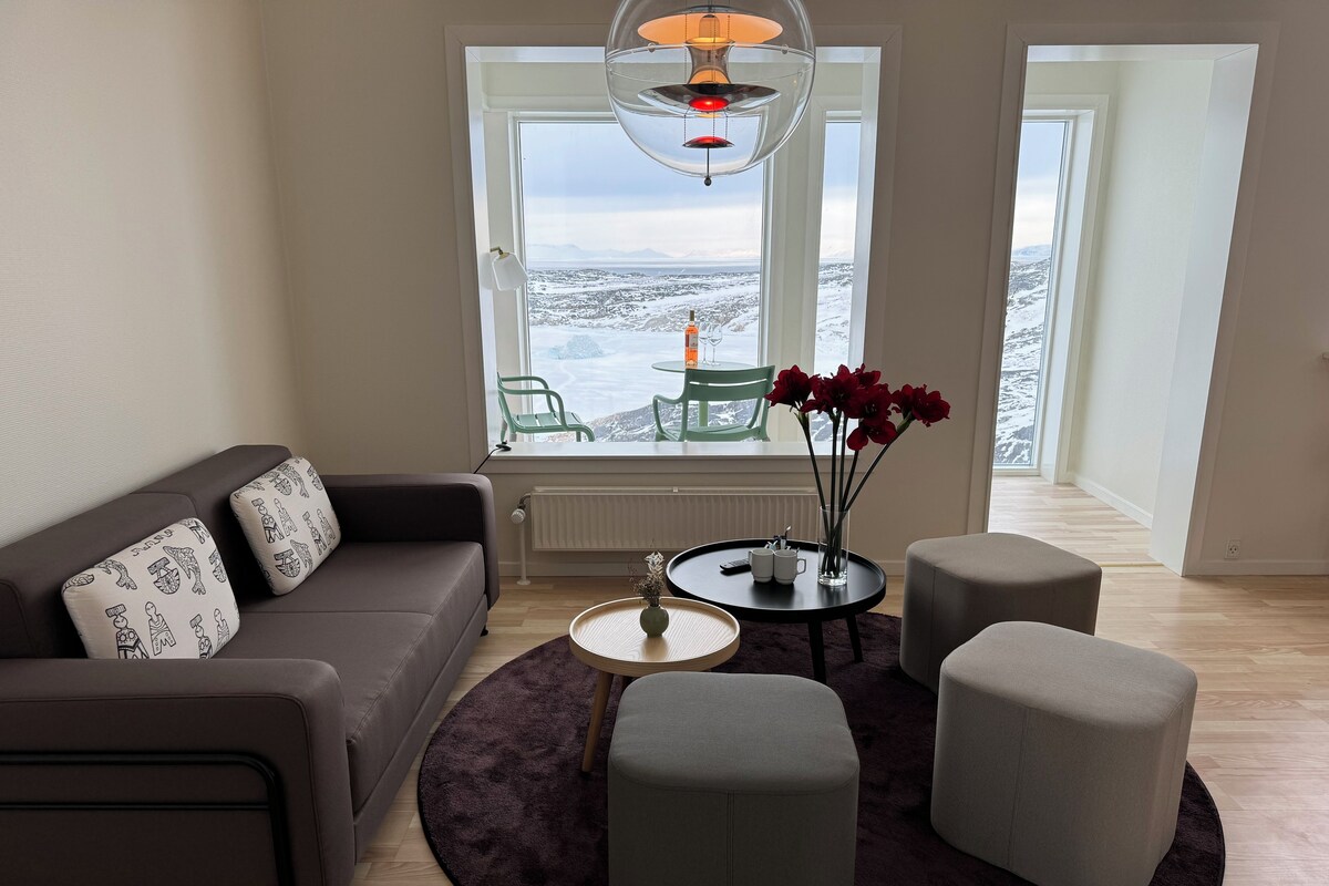 A modern living room with a sofa, ottomans, a coffee table, and a view of a snowy landscape through large windows.