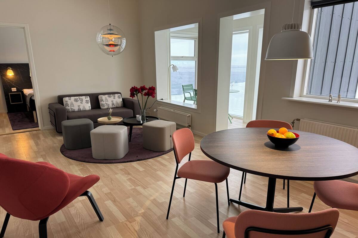 Una sala de estar moderna con una mesa de comedor redonda, sillas rojas, un sofá gris y un florero con flores rojas.