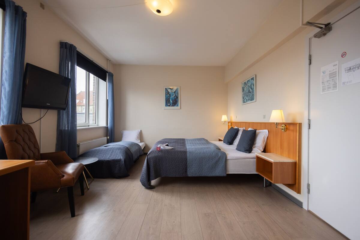 A hotel room with two beds, a desk, a chair, a TV, and artwork on the walls.