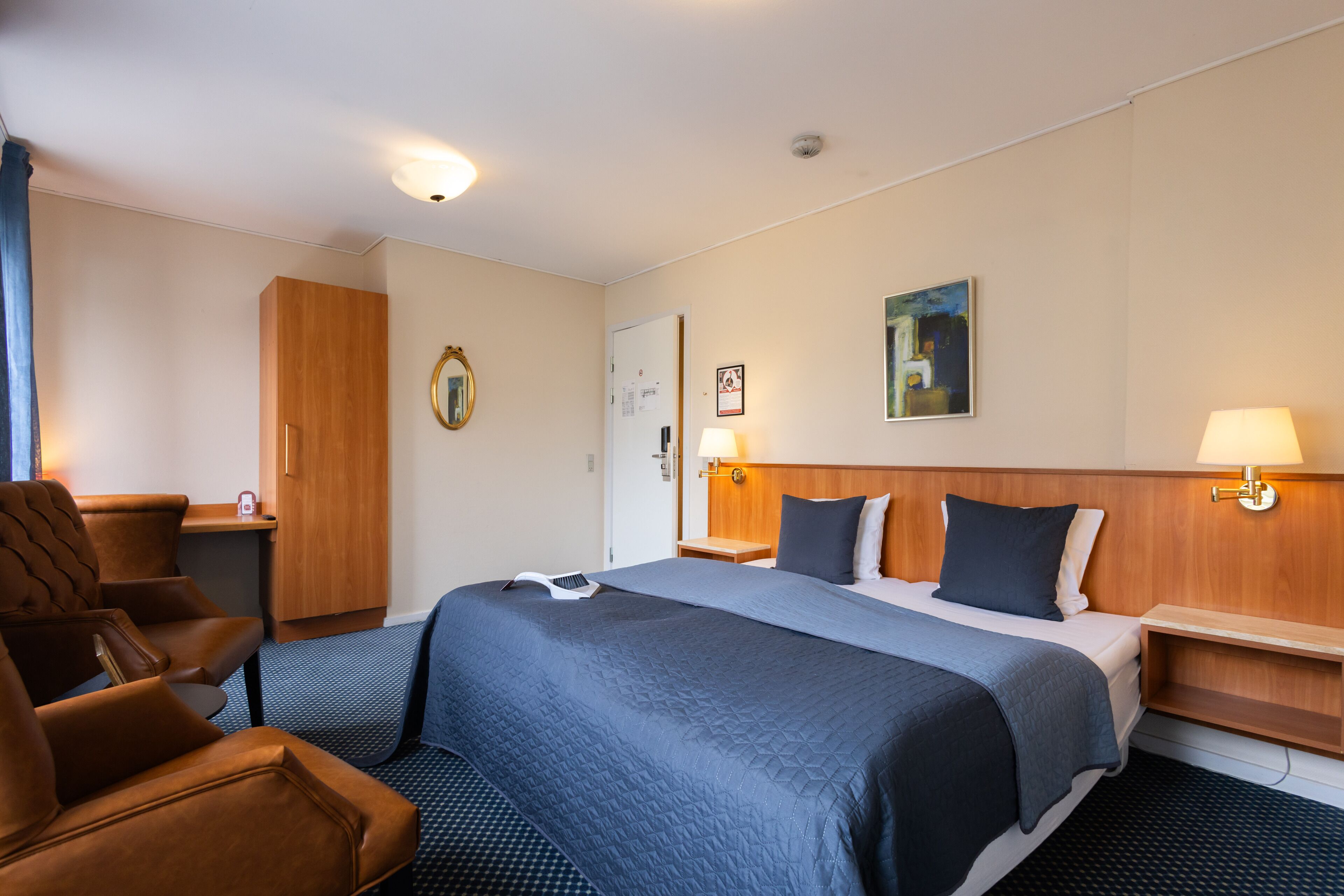 A hotel room with a large bed, a wooden wardrobe, a desk with a chair, and a nightstand with a lamp.