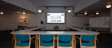 Un salón de conferencias con una larga mesa, sillas azules, una pantalla de proyector y una pizarra.