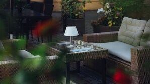 A wicker sofa with a cushion, a small table with a lamp, and chairs in an outdoor seating area with potted plants and a table in the background. - Milling Hotel Gestus (Aalborg)