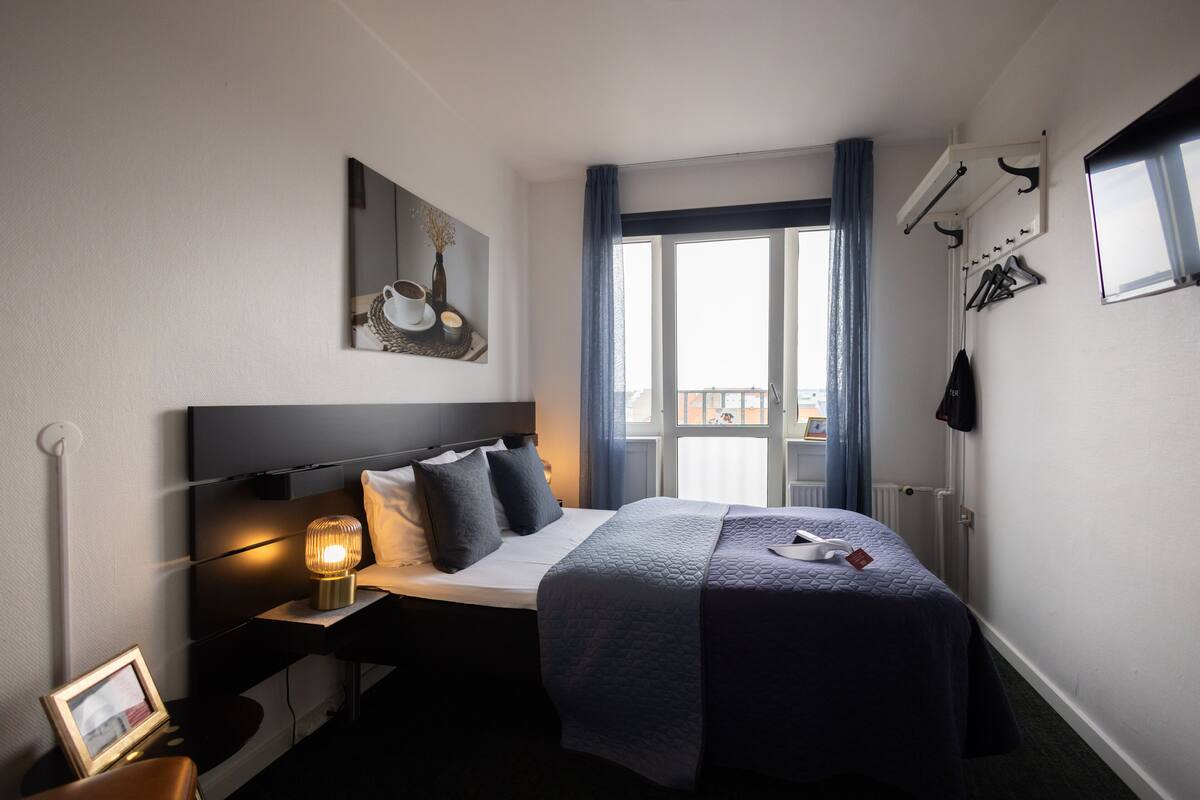 A hotel room with a bed, bedside table, lamp, picture frame, and a window with curtains.