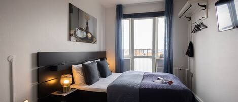 Habitación de hotel con cama, mesita de noche, lámpara, marco de foto y ventana con cortinas.
