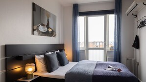 A hotel room with a bed, a bedside lamp, a window with curtains, a coat rack, and a painting on the wall. - Milling Hotel Gestus (Aalborg)