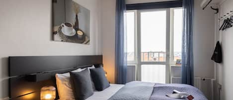 Una habitación de hotel con cama, lámpara de noche, ventana con cortinas, perchero y un cuadro en la pared.