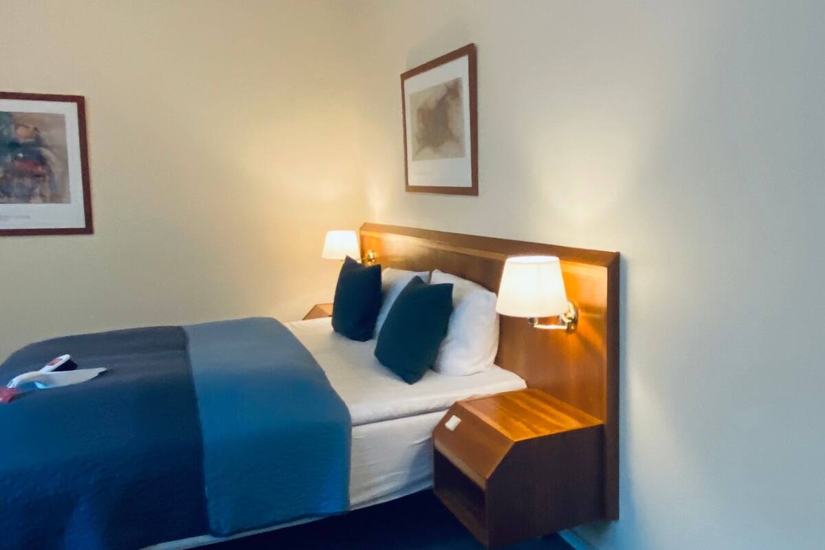 A neatly made bed with a blue blanket, two pillows, and a wooden nightstand with a lamp.