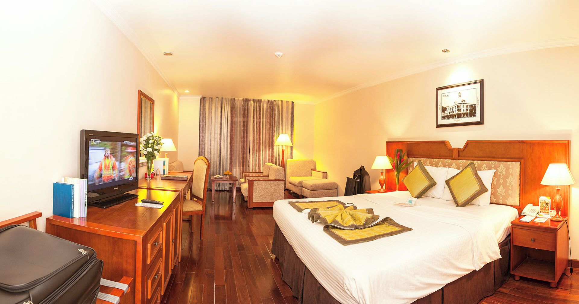A hotel room with a bed, a desk with a television, a chair, a table, and a nightstand with a lamp.