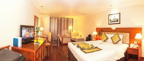 A hotel room with a bed, a desk with a television, a chair, a table, and a nightstand with a lamp.