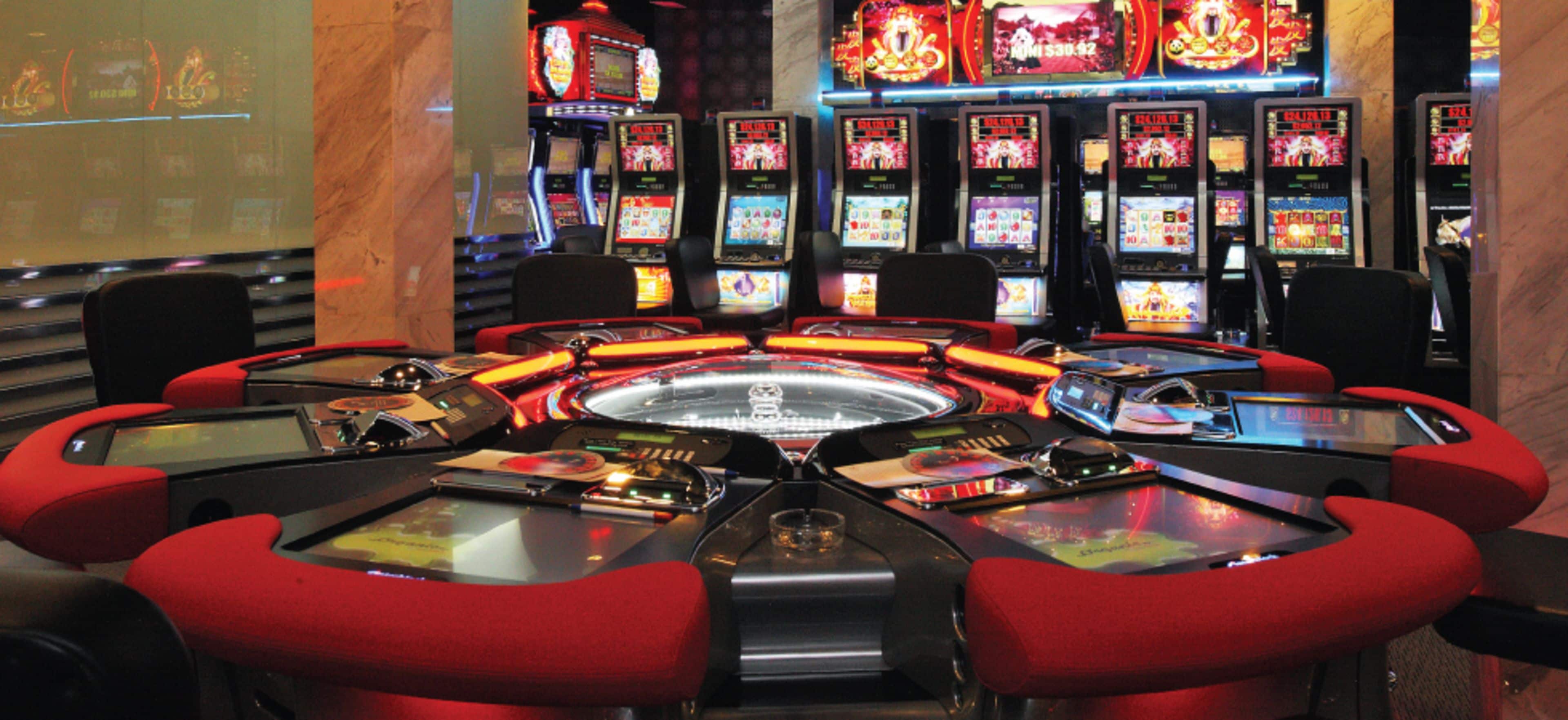 A casino floor with multiple slot machines and red cushioned chairs around a central table.