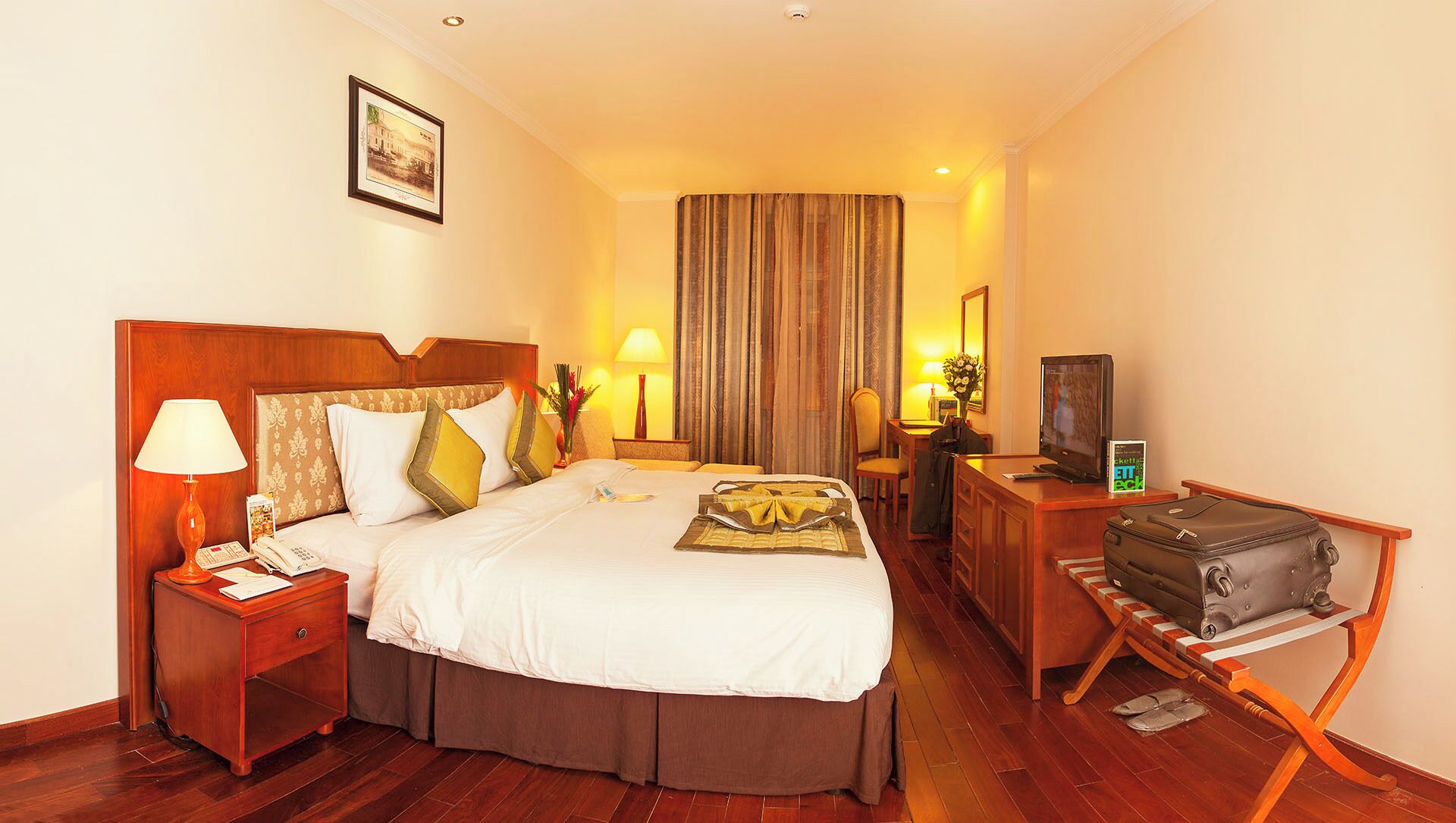 A hotel room with a bed, bedside table, lamp, TV, desk, chair, and a suitcase.
