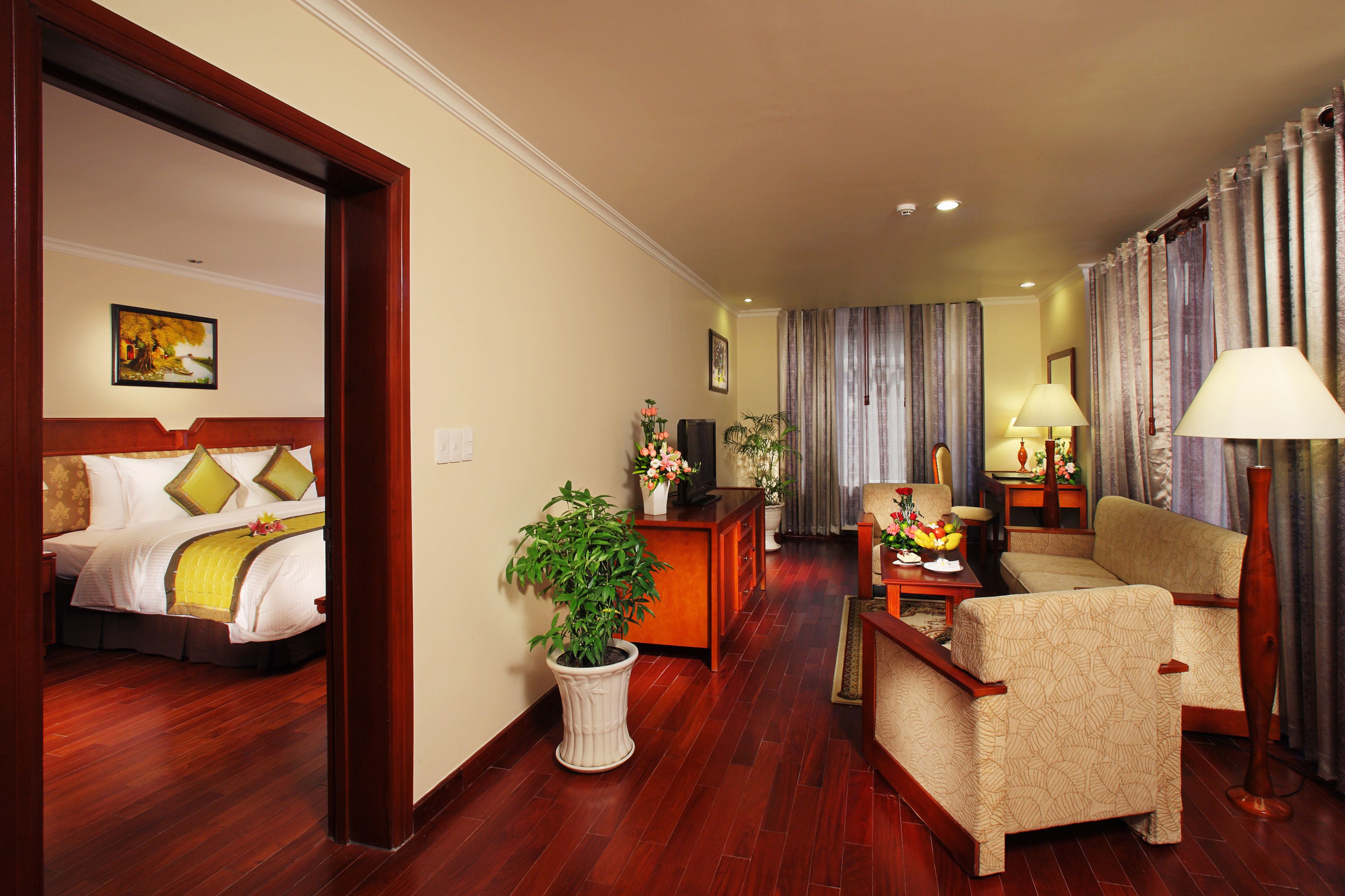 A hotel room with a bed, a sitting area with a sofa and armchairs, a coffee table, a potted plant, and a desk with a lamp.