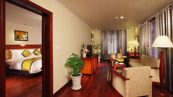 Une chambre d’hôtel comprenant un lit, un coin salon avec un canapé et des fauteuils, une table basse, une plante en pot et un bureau avec une lampe.