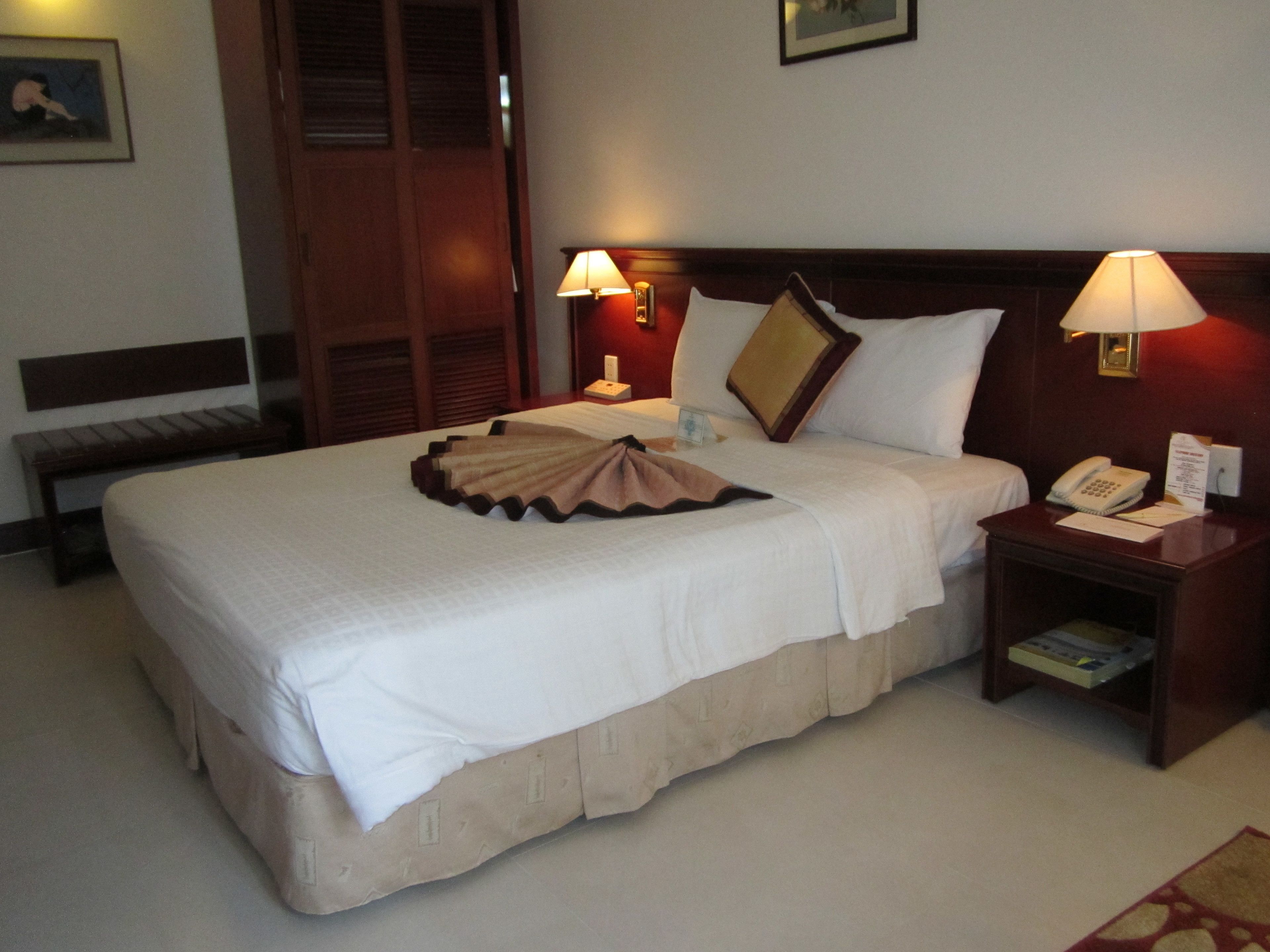 A neatly made bed with a decorative skirt, two bedside lamps, a bedside phone, and a small table with a book and a notepad.