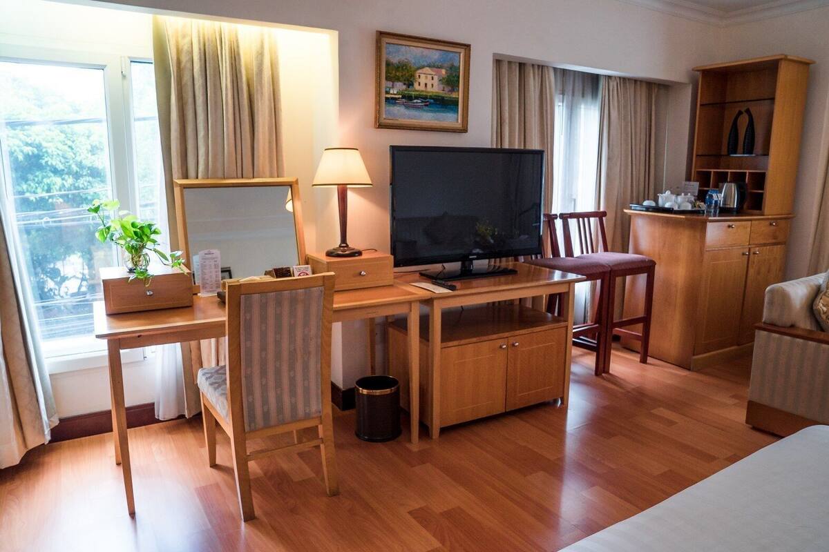 Habitación de hotel con escritorio de madera, televisor, sillón, sofá, mueble pequeño, cesto de basura y ventana con cortinas.
