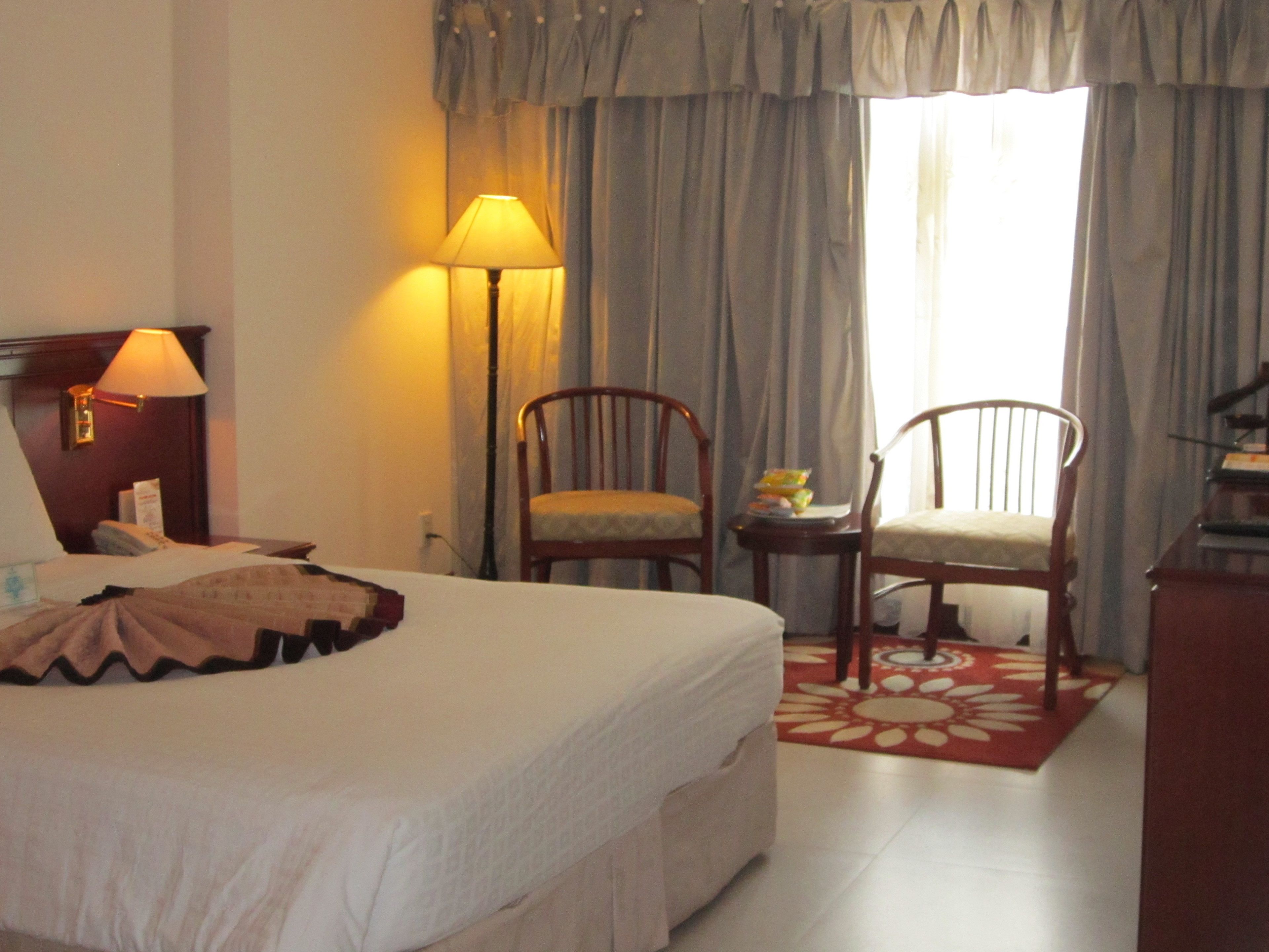 A hotel room with a bed, two chairs, a lamp, and a window with curtains.