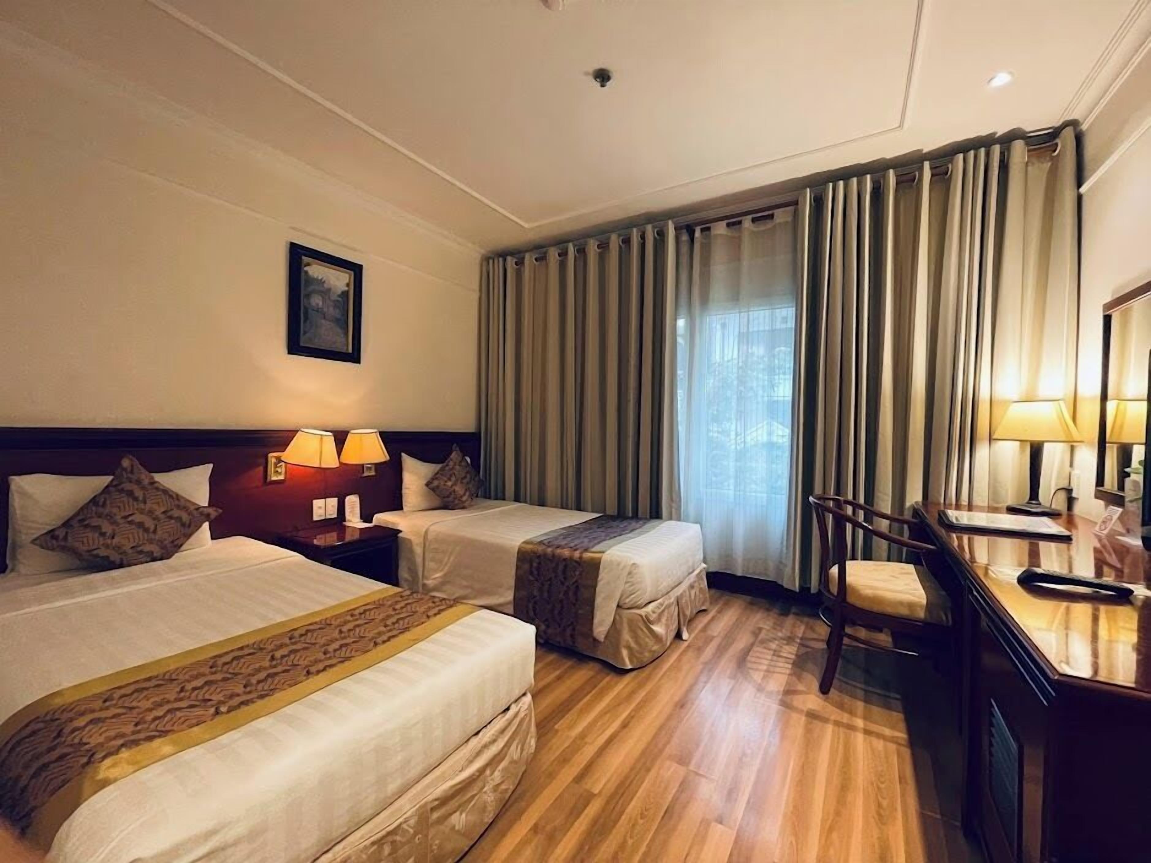 A hotel room with two beds, a desk, and a chair. There is a window with curtains, a painting on the wall, and a lamp on the desk.