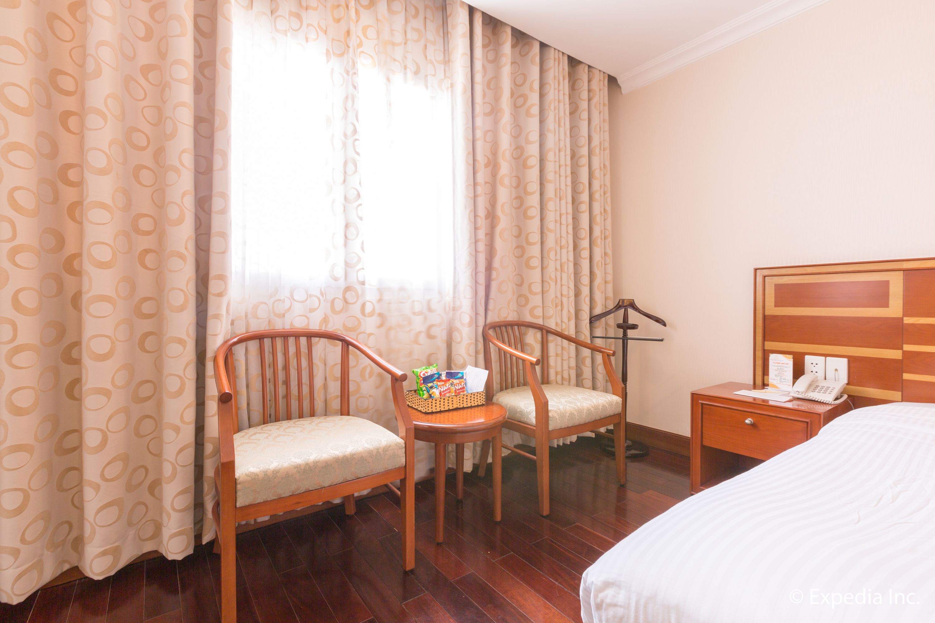 A hotel room with two wooden chairs, a small table with snacks, a bedside table with a phone, and a large bed.