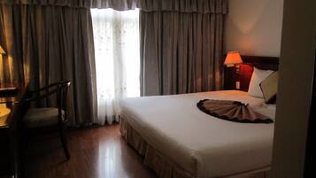 Quarto de hotel com cama, escrivaninha, cadeira, mesa de cabeceira e janela com cortinas.