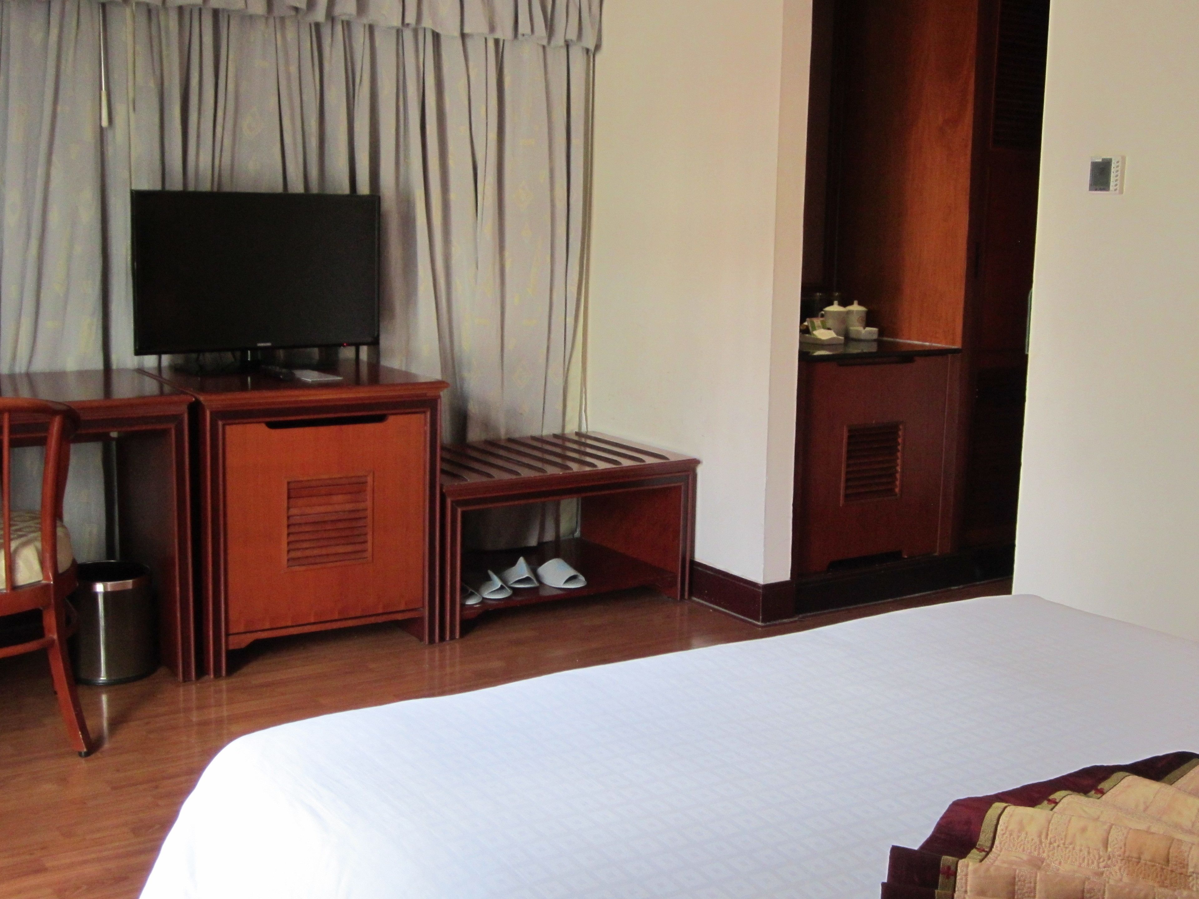 Quarto de hotel com uma cama grande, uma escrivaninha de madeira com televisão e uma pequena prateleira com objetos.
