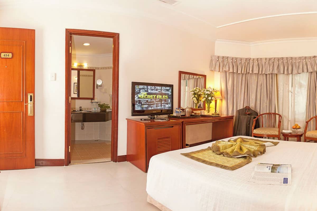 Habitación de hotel con una cama grande, un escritorio con televisor, dos sillas, un espejo y una ventana con cortinas.