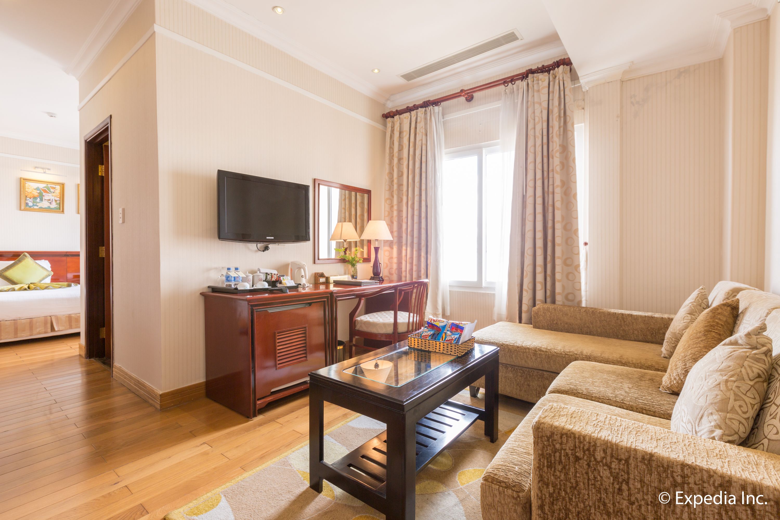 Habitación de hotel con piso de madera, un sofá, un televisor montado en la pared, un escritorio con lámpara y una ventana con cortinas.