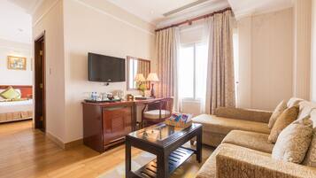 Habitación de hotel con piso de madera, un sofá, un televisor montado en la pared, un escritorio con lámpara y una ventana con cortinas.