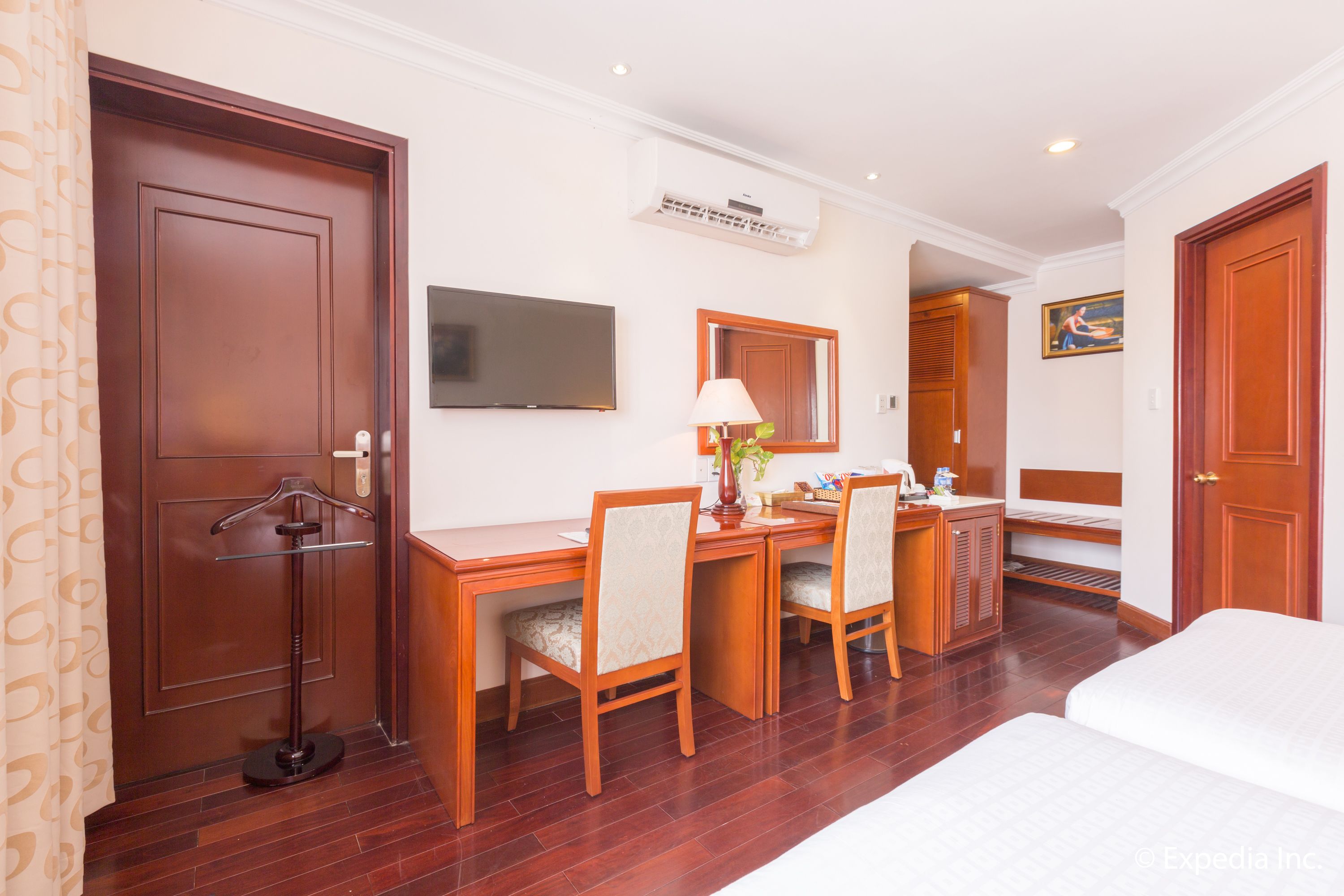 A hotel room with a wooden desk, two chairs, a bed, a TV, and a painting on the wall.