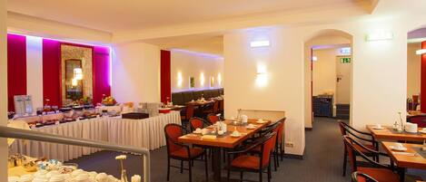 Comedor de hotel con varias mesas y sillas, un bufé con distintos alimentos, y una zona de estar con una decoración de colores rojo y blanco.