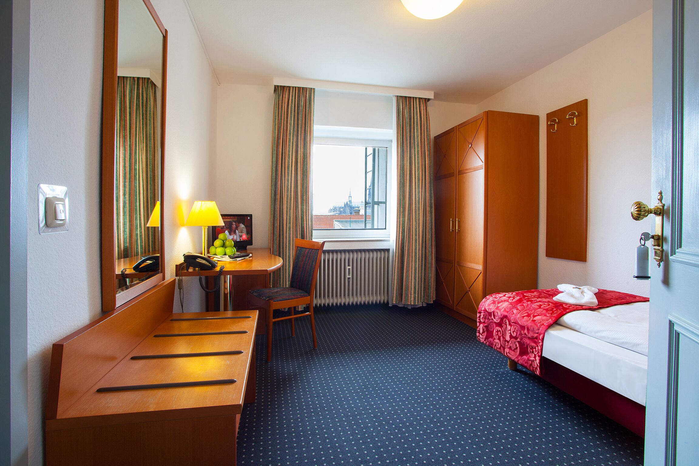 Habitació d'hotel amb un llit, una taula de treball amb una cadira, un mirall, una finestra amb cortines i un radiador.