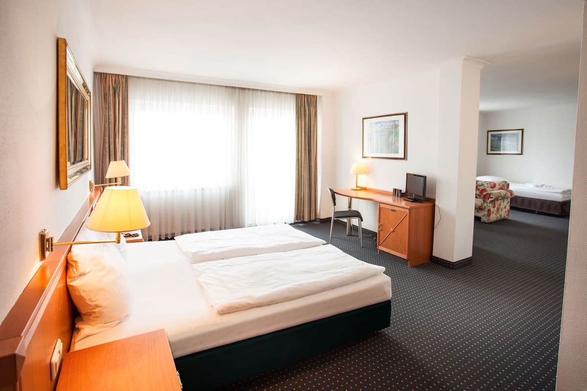 Habitación de hotel con cama doble, escritorio con silla, un pequeño sofá y otra cama al fondo.