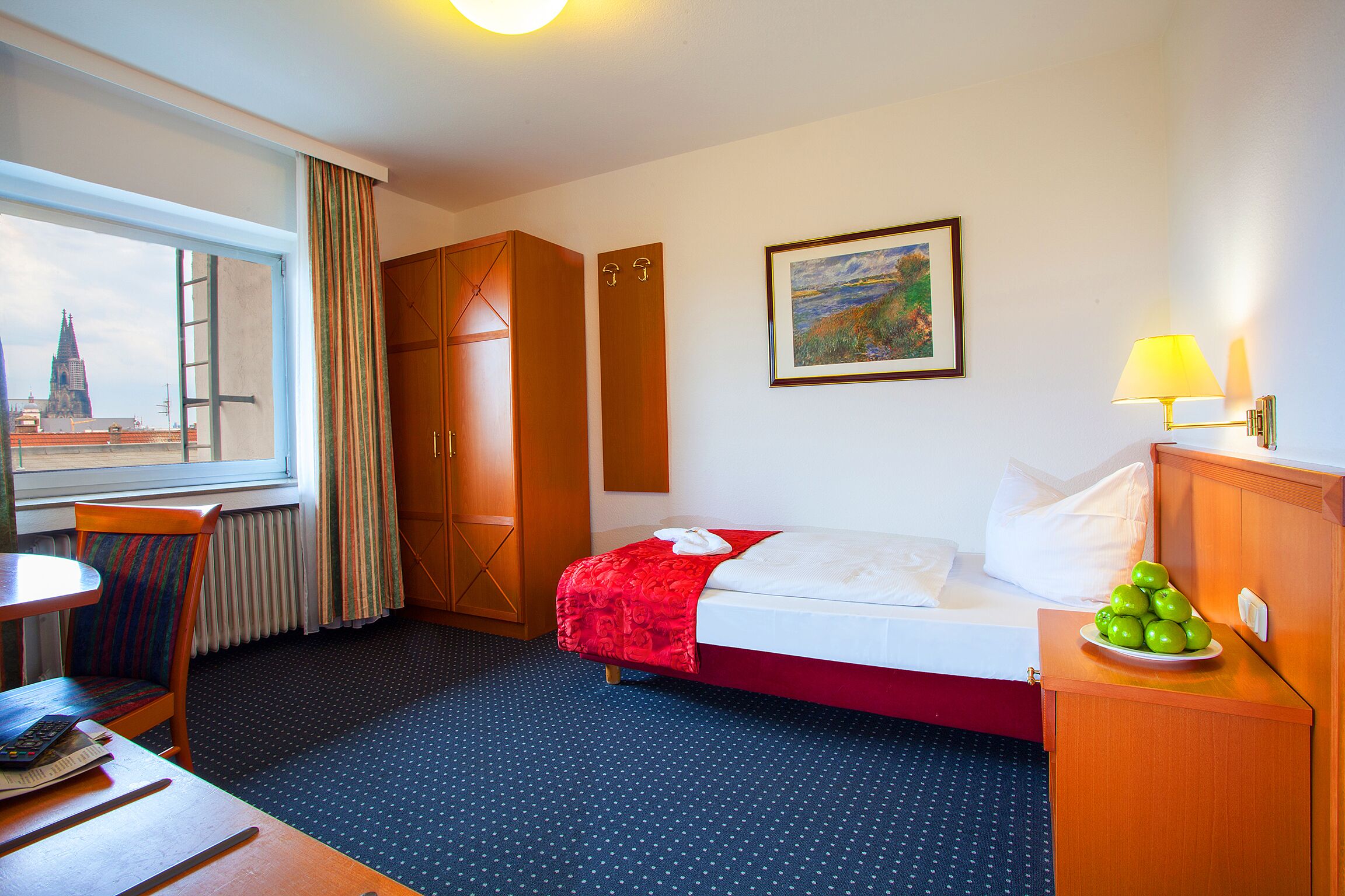 Habitación de hotel con cama, escritorio, silla, ventana con cortinas, un cuadro en la pared y una lámpara en la mesita de noche.
