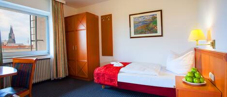 Habitación de hotel con cama, escritorio, silla, ventana con cortinas, un cuadro en la pared y una lámpara en la mesita de noche.