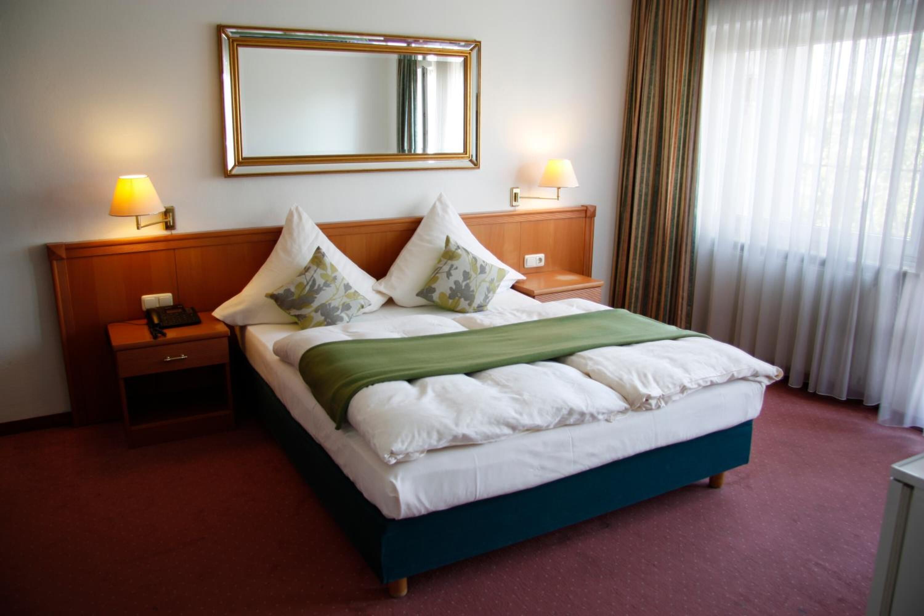Une chambre d’hôtel avec un grand lit, deux tables de chevet, une commode, un miroir et une fenêtre avec des rideaux.