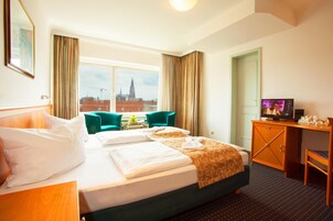 Habitación de hotel con cama, dos sillas, escritorio y televisor, y vista a la ciudad.