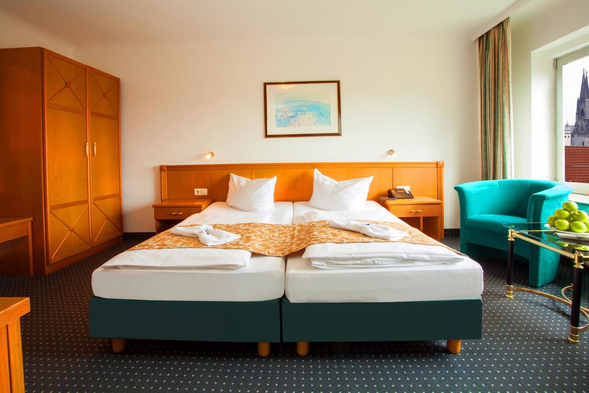 Habitación de hotel con cama doble, armario de madera, sillón azul turquesa y vista a un paisaje urbano por la ventana.