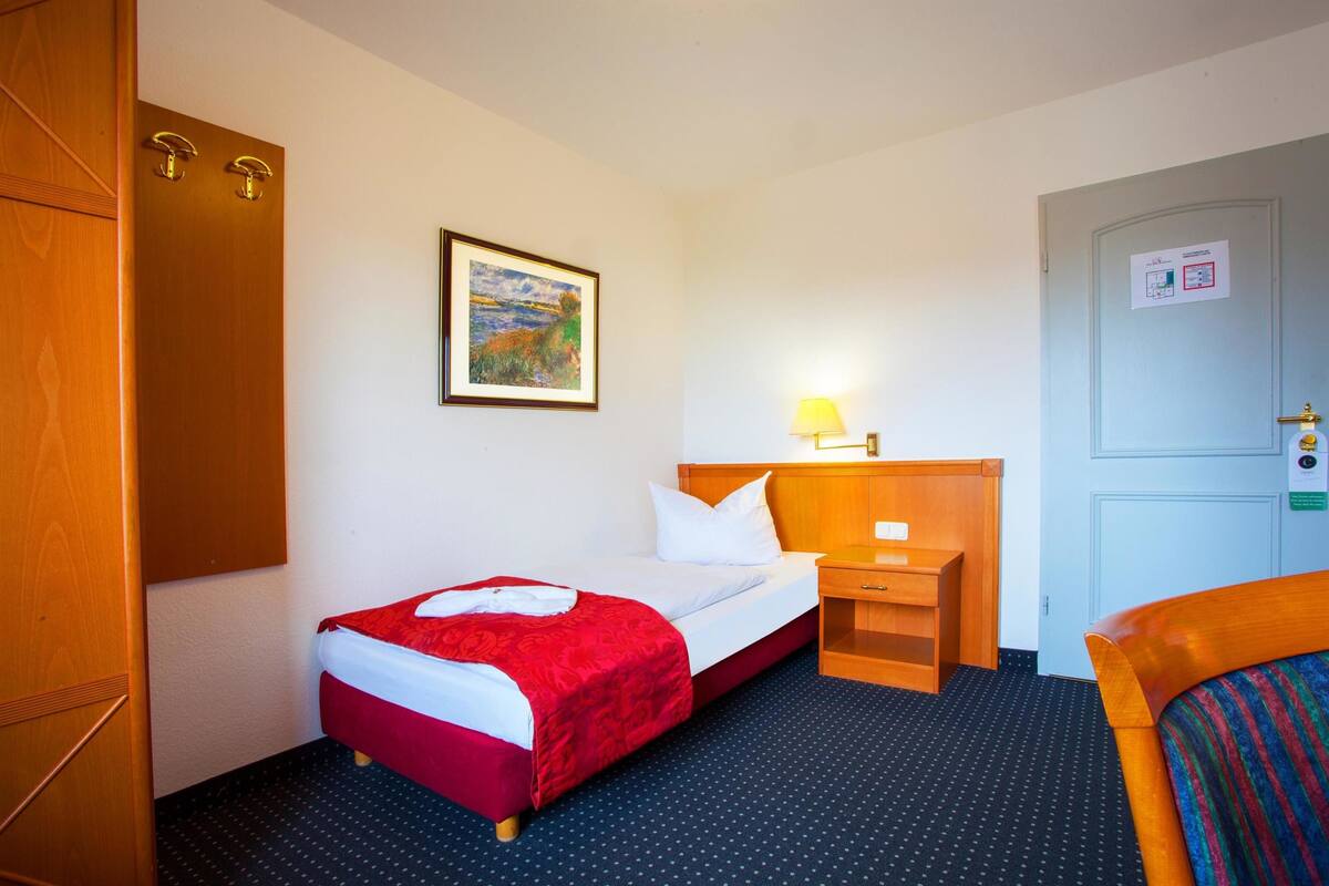 Habitación de hotel con una cama individual, una mesita de noche con lámpara, un armario de madera y un cuadro enmarcado en la pared.