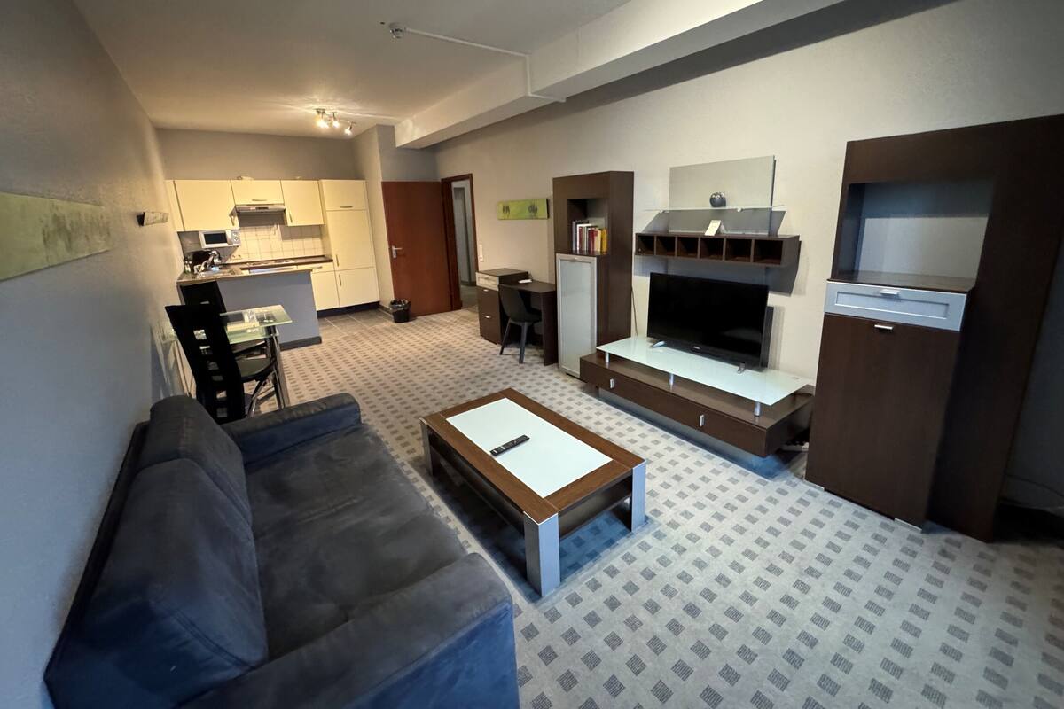 A modern living room with a sofa, a coffee table, a TV, and a kitchen area in the background.