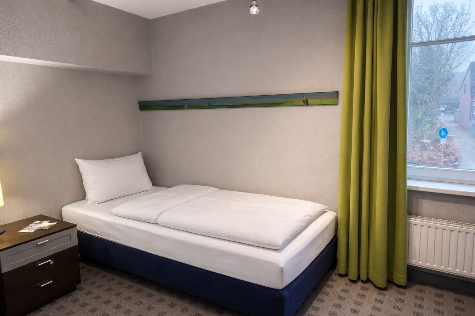 Una cama individual en una habitación con ventana, una mesita de noche y un estante fijado a la pared.