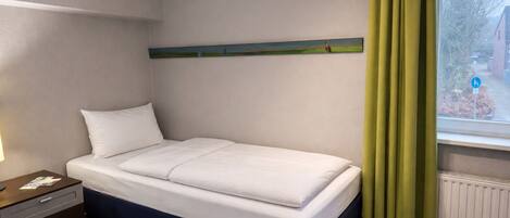 Una cama individual en una habitación con ventana, una mesita de noche y un estante fijado a la pared.
