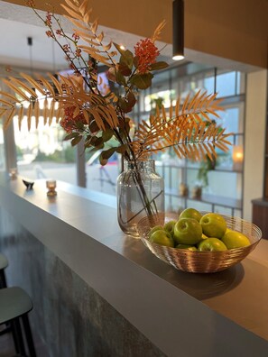 A clear glass vase with dried foliage and red flowers on a countertop, next to a copper wire basket with green apples.